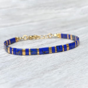 May include: A bracelet featuring alternating rectangular lapis lazuli beads and thin gold-toned metal spacers. The piece includes a gold-toned metal chain extender and a lobster claw clasp, creating a continuous band of deep blue and metallic gold.