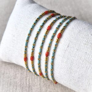 Op de afbeelding: Een set van vijf delicate armbanden met een herhalend patroon van kleine metallic gouden kralen, lichtblauwe turquoise gespikkelde kralen en gladde cilindrische koraalkleurige kralen, gerangschikt in een dun en flexibel strengontwerp.