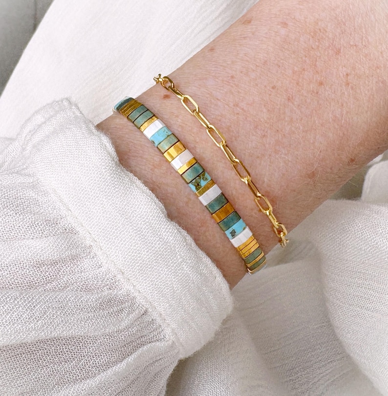 May include: A gold chain bracelet featuring elongated oval links paired with a second bracelet composed of small, rectangular, flat beads in alternating colors of turquoise, white, and metallic gold, arranged in a repeating horizontal pattern.
