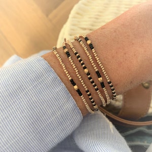Op de afbeelding: Een set van vijf gouden en zwarte kralen armbanden gestapeld op een pols. De armbanden zijn gemaakt van kleine, ronde kralen met een subtiele glans.