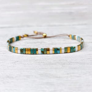 May include: A delicate bracelet featuring a repeating pattern of rectangular turquoise, metallic gold, and silver beads. The bracelet is constructed with a thin brown cord and includes an adjustable sliding closure with two small gold beads.