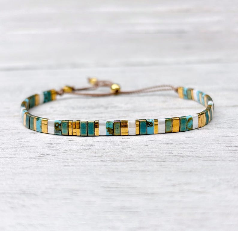 May include: A delicate bracelet featuring a repeating pattern of rectangular turquoise, metallic gold, and silver beads. The bracelet is constructed with a thin brown cord and includes an adjustable sliding closure with two small gold beads.