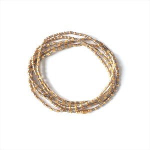 Könnte beinhalten: Ein mehrsträngiges Armband aus kleinen, zylindrischen Perlen in abwechselnden metallischen Gold- und matten Grautönen. Die Perlen sind in einer kreisförmigen Anordnung aufgereiht und erzeugen ein geschichtetes Aussehen mit einem einheitlichen Farbmuster.