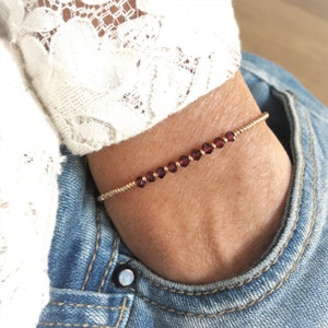 Può includere: Un braccialetto dorato con piccole pietre preziose rotonde di colore rosso scuro. Il braccialetto è indossato su un polso, sullo sfondo di jeans e un top di pizzo bianco. Le pietre preziose sono uniformemente distanziate lungo la fascia dorata.