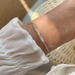 Könnte beinhalten: Ein zartes Armband mit kleinen, facettierten, durchscheinenden hellgrauen Perlen, die mit winzigen, polierten goldfarbenen Metallkugeln durchsetzt sind. Das Armband ist auf einer dünnen Schnur aufgezogen und liegt auf der Haut.