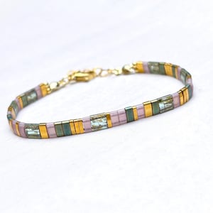 Op de afbeelding: Een armband bestaande uit afwisselende rechthoekige kralen in metallic goud, mat lichtroze en iriserend groenblauw. De armband is voorzien van een goudkleurige metalen karabijnsluiting en een verstelbare kettingverlenging aan één uiteinde.