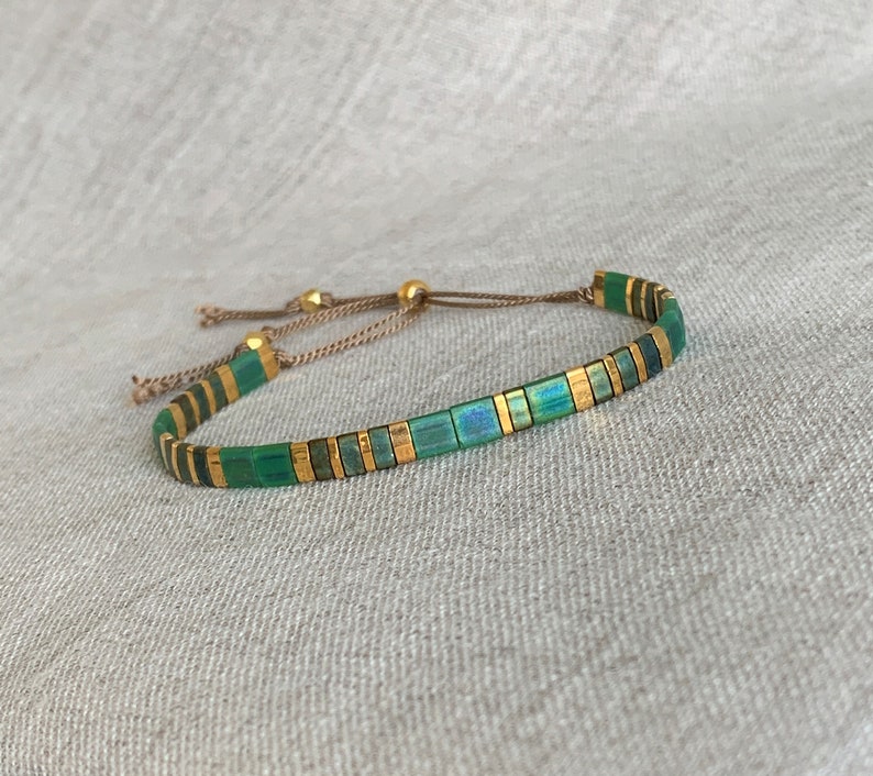 Puede incluir: Un brazalete de cuentas verdes y doradas con cierre de cord&oacute;n marr&oacute;n. Las cuentas son cuadradas y tienen un sutil brillo iridiscente.