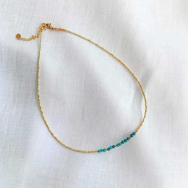 May include: A delicate beaded necklace featuring a strand of small cream-colored seed beads interspersed with metallic gold beads. A section of the necklace includes a series of round, vibrant turquoise-colored beads. It has a gold-tone metal clasp.