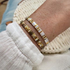Könnte beinhalten: Zwei gestapelte Armbänder aus rechteckigen Perlen. Ein Armband besteht aus weißen, goldenen und braun marmorierten Perlen. Das zweite Armband besteht aus abwechselnden weißen und goldenen Perlen. Beide Armbänder werden am Handgelenk getragen.