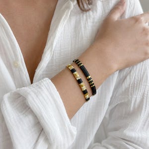 Op de afbeelding: Twee kralenarmbanden gedragen om de pols. De ene armband heeft afwisselend matzwarte en metallic gouden rechthoekige kralen. De tweede armband bestaat uit matzwarte kralen afgewisseld met secties van goud en wit patroon kralen.