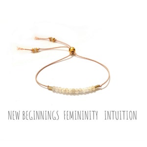 May include: A delicate bracelet featuring small, light-colored beads strung on a thin, adjustable cord. The bracelet has gold-colored metal accents and a sliding bead closure. The words "NEW BEGINNINGS FEMININITY INTUITION" are printed below the bracelet.