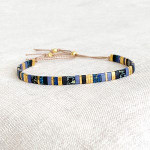 Peut inclure: Bracelet réglable avec un motif mosaïque. Il présente des perles rectangulaires dorées, noires, bleues et à motifs. Le bracelet est monté sur un cordon beige avec des accents dorés. Le bracelet est présenté sur une surface blanche.