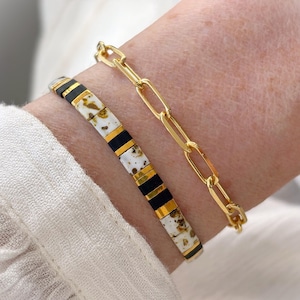 May include: A set of two bracelets. One bracelet features a gold-tone chain with elongated oval links. The second bracelet consists of flat, rectangular beads in a repeating pattern of matte black, metallic gold, and white with brown speckles.