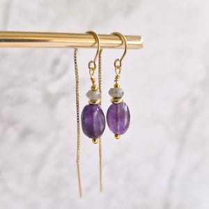 Peut inclure: Une paire de boucles d'oreilles pendantes en métal doré avec une perle d'améthyste violette, une petite perle en pierre grise et des intercalaires dorés. Les boucles sont suspendues à une fine chaîne dorée qui traverse le lobe de l'oreille.