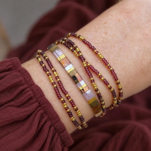 May include: A multi-strand bracelet featuring a central row of rectangular tiles in shades of metallic gold, bronze, copper, and soft lavender, flanked by several delicate strands of small, round, deep burgundy and metallic gold seed beads.