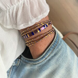 May include: A multi-strand wrap bracelet featuring small matte taupe seed beads, metallic gold spacer beads, and a central row of rectangular tiles in alternating deep blue, purple, and metallic gold colors, creating a layered appearance.