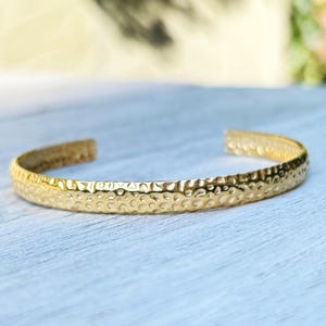 Pode incluir: Uma pulseira tipo bracelete de tom dourado com uma superfície texturizada e martelada, apresentando um design aberto na parte traseira. A banda de metal tem um acabamento reflexivo e largura constante com pequenas reentrâncias irregulares.