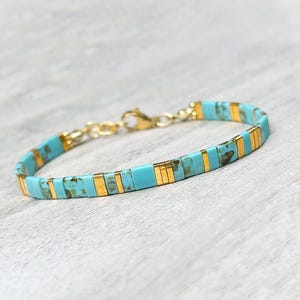Op de afbeelding: Een armband met afwisselende rechthoekige turquoise kralen met bruine marmering en dunne, gepolijste goudkleurige metalen tussenstukjes. Het sieraad wordt vastgemaakt met een goudkleurige metalen karabijnsluiting en een korte bijpassende ketting.
