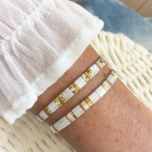 Peut inclure: Deux bracelets composés d'un motif répétitif de petites perles rectangulaires blanches et d'entretoises en or métallique. Les perles ont une finition lisse et brillante, et les bracelets sont portés ensemble au poignet pour un effet superposé.