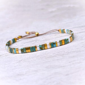 May include: A delicate bracelet featuring a repeating pattern of rectangular beads in turquoise, metallic gold, and white. The bracelet is constructed with a thin brown cord and includes a sliding knot closure mechanism for adjustable sizing.