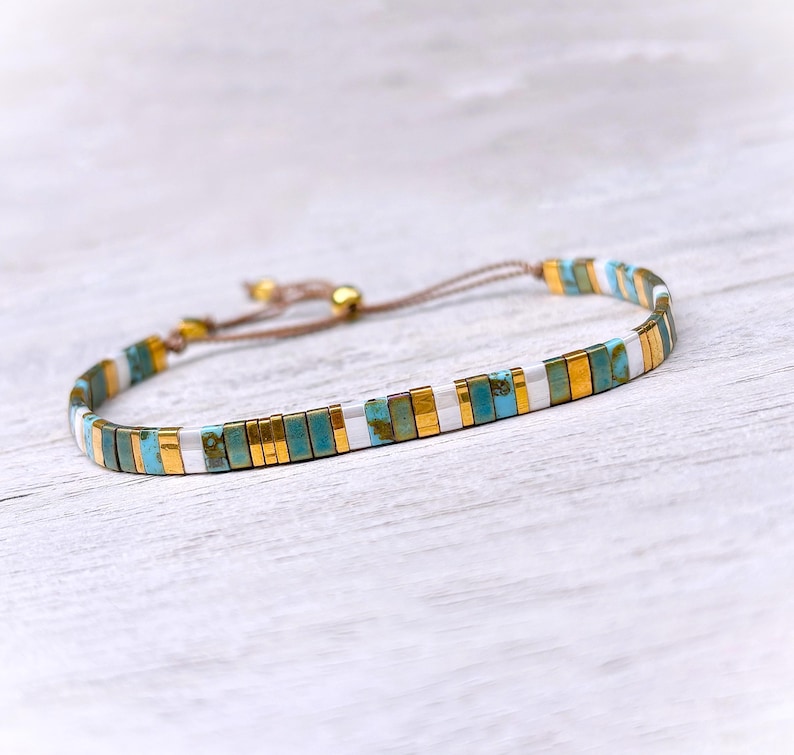 May include: A delicate bracelet featuring a repeating pattern of rectangular beads in turquoise, metallic gold, and white. The bracelet is constructed with a thin brown cord and includes a sliding knot closure mechanism for adjustable sizing.