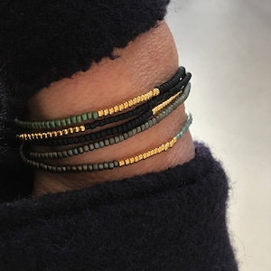 Könnte beinhalten: Ein Stapel Perlenarmbänder in Schwarz, Grün und Gold. Die Armbänder sind übereinander am Handgelenk einer Person gelegt.