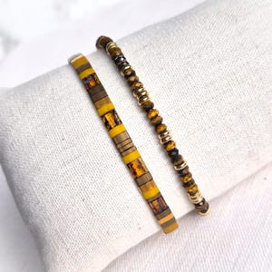 Peut inclure: Deux bracelets. L'un est de forme rectangulaire plate avec des segments jaunes et bruns. L'autre est un bracelet de perles avec des perles marron et dorées. Les deux bracelets sont sur une surface blanche.