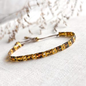 Peut inclure: Un bracelet délicat composé de perles rectangulaires couleur ambre alternées avec de petites entretoises métalliques dorées. Le design comprend un cordon fin marron avec un fermoir coulissant réglable, offrant une allure raffinée et structurée.