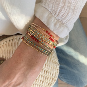 Op de afbeelding: Een stapel van meerdere dunne kralenarmbanden met een verscheidenheid aan kleuren, waaronder metallic goud, turkoois, koper en felrood. De armbanden zijn gemaakt van kleine cilindrische en ronde kralen die in terugkerende patronen zijn gerangschikt.