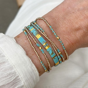 Op de afbeelding: Een armbandset met meerdere strengen, bestaande uit kleine metallic goudkleurige kralen en platte rechthoekige turquoise kralen met bruine aders. Het ontwerp bevat afwisselende segmenten van goudkleurige tussenstukjes tussen de turquoise kralen.