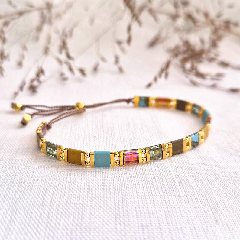 May include: A delicate bracelet featuring a series of alternating rectangular beads in shades of turquoise, brown, and iridescent pink, separated by small gold-toned metallic spheres. The bracelet is secured with a brown adjustable cord closure.