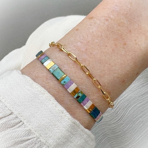 Può includere: Due braccialetti su un polso. Uno è un braccialetto a catena d'oro. L'altro è un braccialetto di perline con perline rettangolari nei toni del verde acqua, viola, bianco, oro e blu scuro.