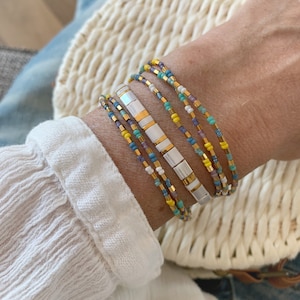 May include: A multi-strand bracelet set featuring a central row of rectangular white and gold-tone beads flanked by thinner strands of small seed beads in shades of yellow, turquoise, blue, white, and metallic gold, creating a layered look.
