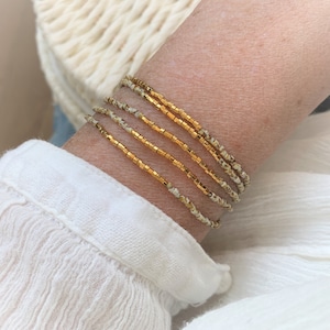 Könnte beinhalten: Ein Set aus vier zierlichen Armbändern, bestehend aus einer Kombination von metallisch goldenen quadratischen Perlen und kleinen, gesprenkelten cremeweißen Perlen. Die Armbänder sind gestapelt und zeigen ein Muster aus goldenen und cremefarbenen Perlen.