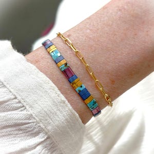 Op de afbeelding: Een tweedelige armbandset bestaande uit een goudkleurige schakelarmband en een kralenarmband samengesteld uit kleine, platte, rechthoekige tegeltjes in tinten turquoise, magenta, blauw en metallic goud in een herhalend patroon.