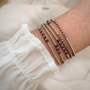 Peut inclure: Un ensemble de cinq bracelets délicats avec de petites perles dorées et des perles de pierres précieuses violettes foncées. Les bracelets sont empilés sur le poignet d'une personne.