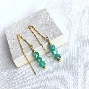 Könnte beinhalten: Ein Paar filigrane Ohrringe. Jeder Ohrring hat drei quadratische, hellgrüne Perlen und eine kleine goldene Perle, die an einer feinen Goldkette aufgereiht sind. Die Ohrringe werden auf einer weißen Oberfläche präsentiert.