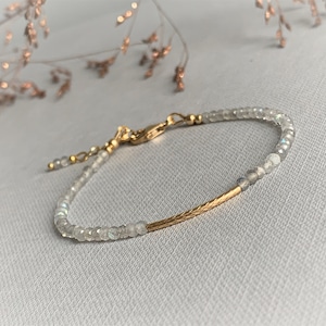 Può includere: Un delicato braccialetto d'oro con una sottile barra dorata e piccole perline bianche iridescenti.