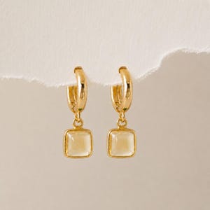 Könnte beinhalten: Ein Paar goldene Creolen, die jeweils mit einem kleinen, quadratischen, durchscheinenden hellgelben Steinanhänger versehen sind. Die Ohrringe haben eine polierte metallische Oberfläche und ein schlichtes Design mit einem sicheren Klappverschluss.