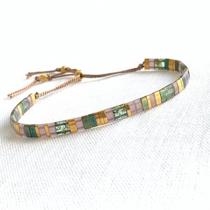 Può includere: Un braccialetto delicato realizzato con piccole perline colorate in tonalità rosa, verde e oro. Il braccialetto è infilato su un cordoncino sottile e ha una chiusura semplice e regolabile.