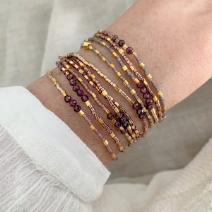 Può includere: Un set di braccialetti di perline in tonalità marrone, oro e viola. I braccialetti sono realizzati con perline piccole e presentano alcune perline viola più grandi e sfaccettate.