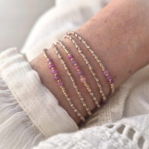 May include: A set of four delicate beaded bracelets featuring small metallic gold beads interspersed with tiny faceted pink gemstones. The bracelets are arranged in a stack, showcasing a repeating pattern of gold and pink beads on a thin cord.