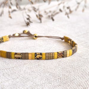Peut inclure: Un bracelet avec fermeture coulissante. Il présente des perles rectangulaires jaunes, dorées et marron. Les perles sont enfilées sur un cordon marron. Le bracelet est présenté sur une surface blanche.