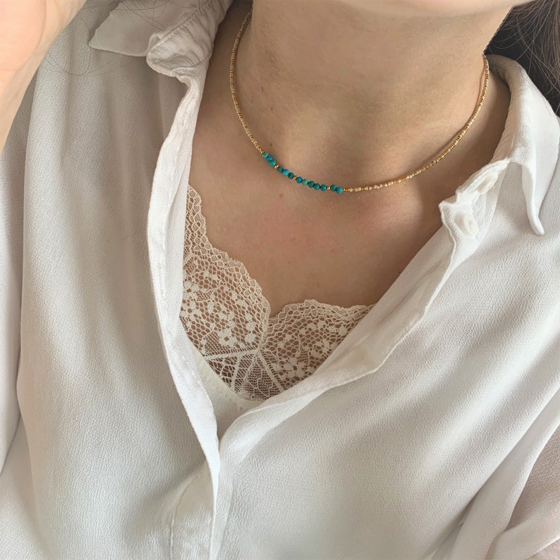 May include: A delicate beaded choker necklace featuring a strand of small metallic gold-toned beads interspersed with a short, asymmetrical section of vibrant turquoise blue beads, resting against the neck in a simple, circular design.