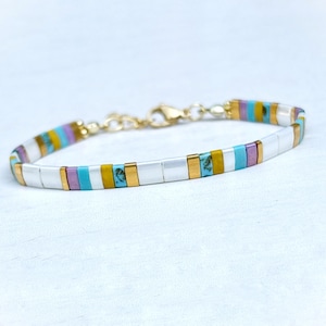 Peut inclure: Un bracelet composé d'un motif répétitif de perles rectangulaires blanches, jaune moutarde, turquoise avec des veines brunes et lavande, séparées par de petites entretoises en métal doré. Le bracelet possède un fermoir mousqueton doré.