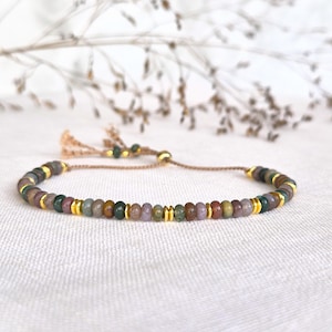 Op de afbeelding: Een kralenarmband met kleine, ronde, aardekleurige stenen in tinten groen, bruin en mauve, afgewisseld met dunne, gepolijste goudkleurige metalen tussenstukjes. De armband heeft een beige koord met een verstelbare schuifsluiting.