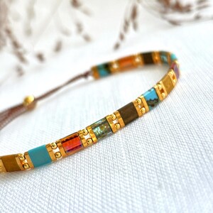 May include: A delicate bracelet featuring a series of rectangular beads in vibrant shades of turquoise, amber, and brown, separated by small gold-toned metallic spacers. The beads are strung on a thin brown cord with an adjustable sliding closure.