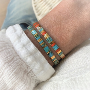 Può includere: Due braccialetti sovrapposti realizzati con piccole perline rettangolari. Le perline presentano un mix di blu turchese con venature marroni, arancione brillante solido e accenti in oro metallizzato, disposti in un motivo lineare su un polso.