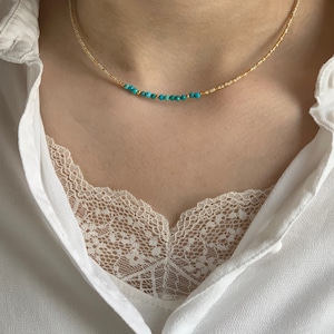 May include: A delicate beaded necklace featuring a strand of small, metallic gold-toned beads interspersed with a central row of vibrant turquoise-colored round beads. The necklace rests against the neck, showcasing a simple and refined design.