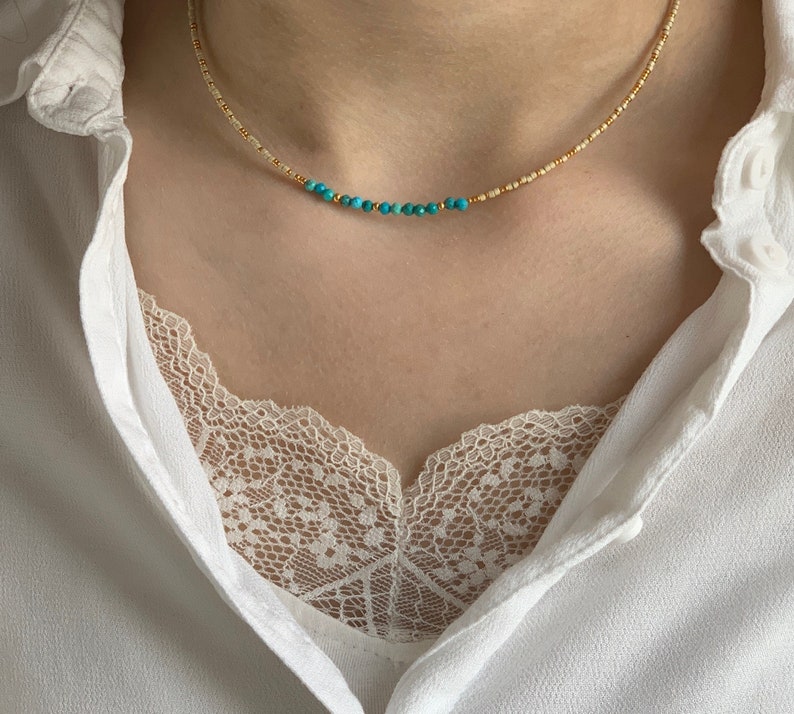 May include: A delicate beaded necklace featuring a strand of small, metallic gold-toned beads interspersed with a central row of vibrant turquoise-colored round beads. The necklace rests against the neck, showcasing a simple and refined design.